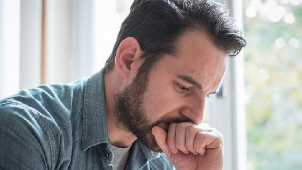 A worried, bearded man rests his hand on his mouth, looking down.