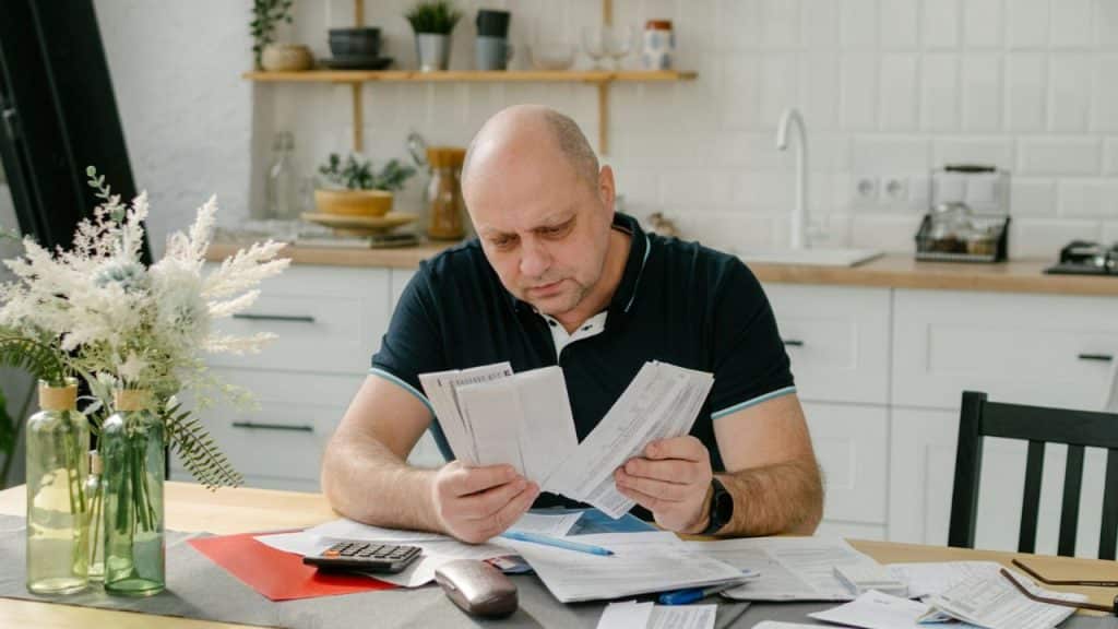 A worried, bald man sits at a kitchen table, reviewing many paper bills.
