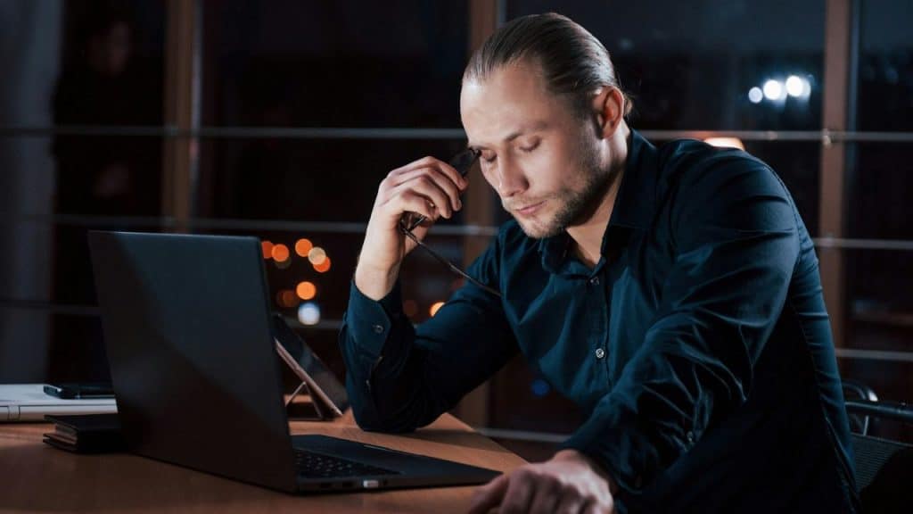 A man tiredly rubs his eyes while working late on a laptop.
