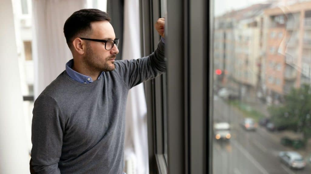 A contemplative man with glasses looks out a large window at a city street.