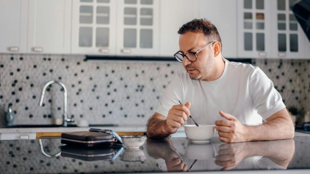 A pensive man with glasses sits at a kitchen counter, stirring a bowl.