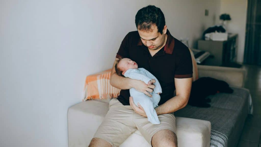 A tired father sits and cradles his newborn baby.