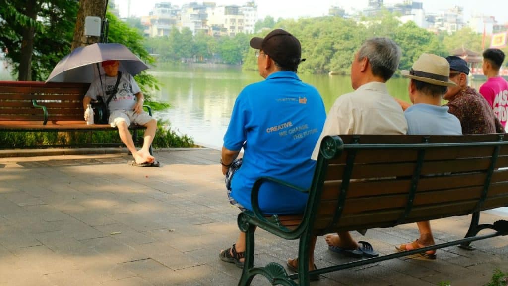 Several middle-aged and older men sit on benches by a lake, looking out at the water.