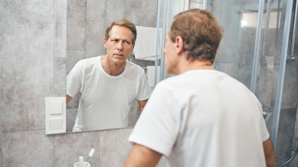 A middle-aged man in a white t-shirt looks seriously at his reflection in a bathroom mirror.