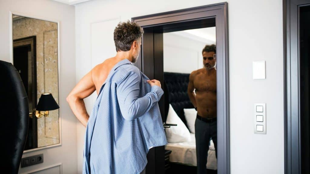 A shirtless, mature man looks at his reflection while putting on a shirt.