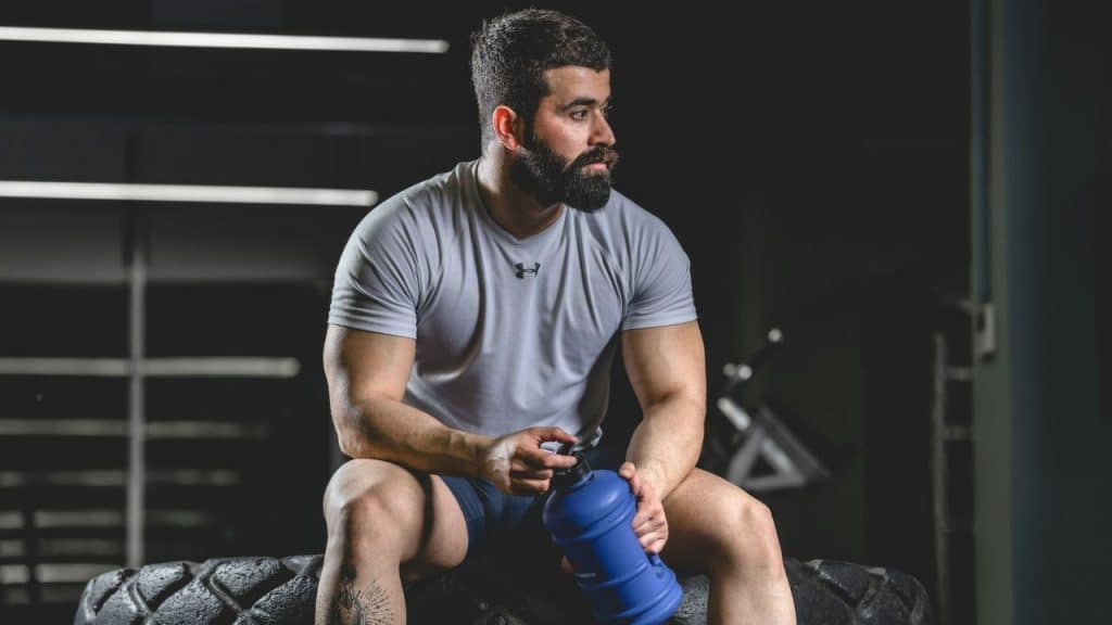 A muscular, bearded man sits on a tire, holding a large water bottle in a gym.