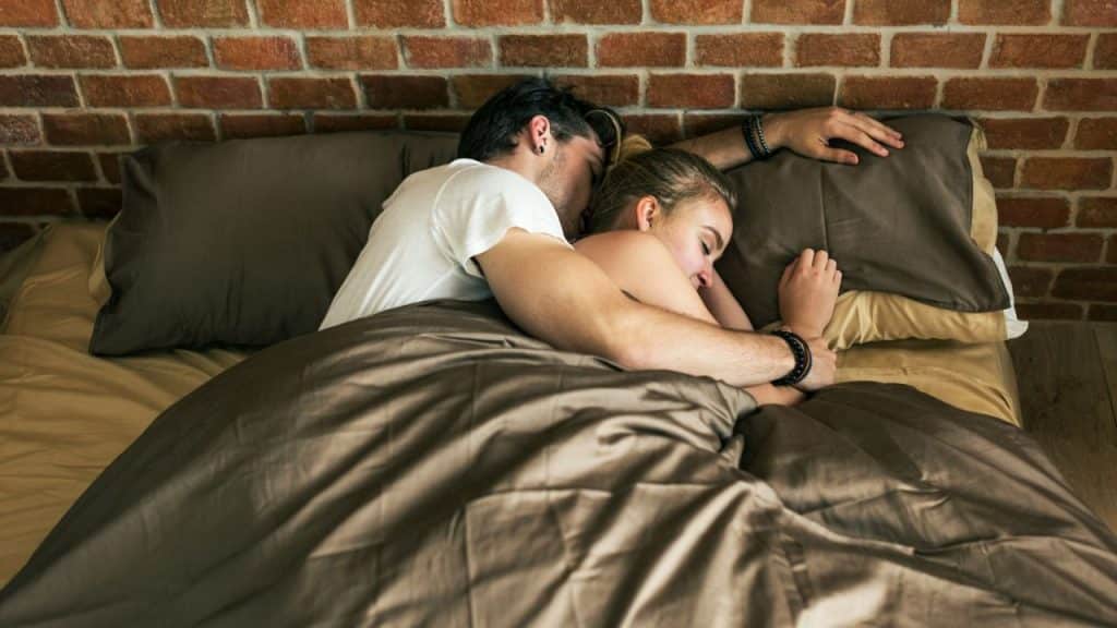 A man and a woman cuddle in bed under a brown blanket against a brick wall.