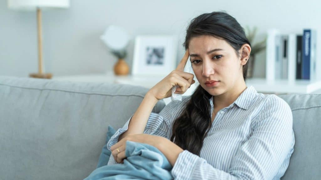 Thoughtful woman sits on a sofa, hand near her forehead, looking troubled.