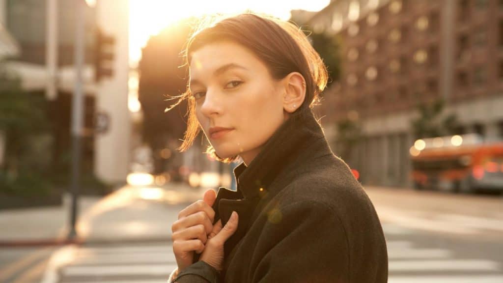 Woman in a dark coat standing on a city street at sunset, looking toward the camera.