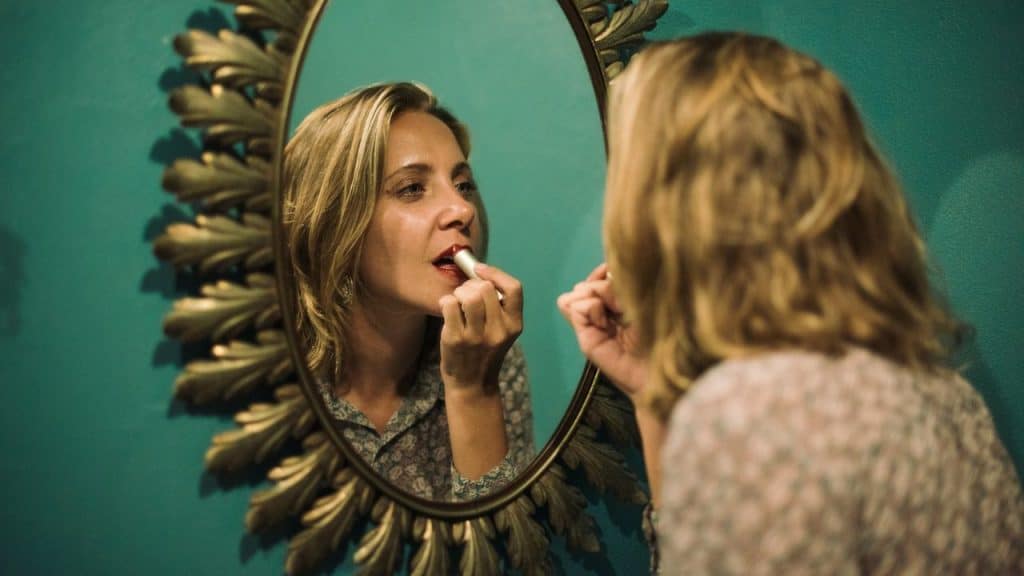 Woman applies red lipstick while looking into a gold-framed mirror on a teal wall.