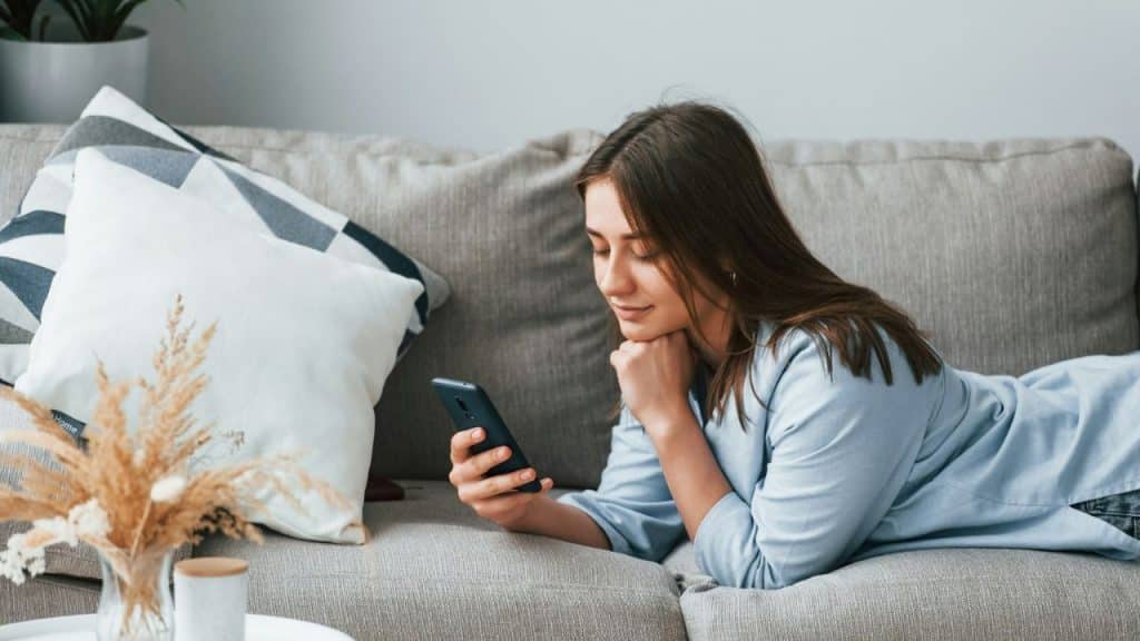 Young woman in a light blue shirt is lying on a sofa, looking at her phone.