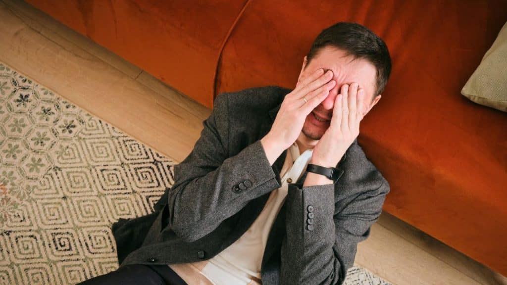 Man in a jacket lying on the floor covers his face with both hands, looking distraught.