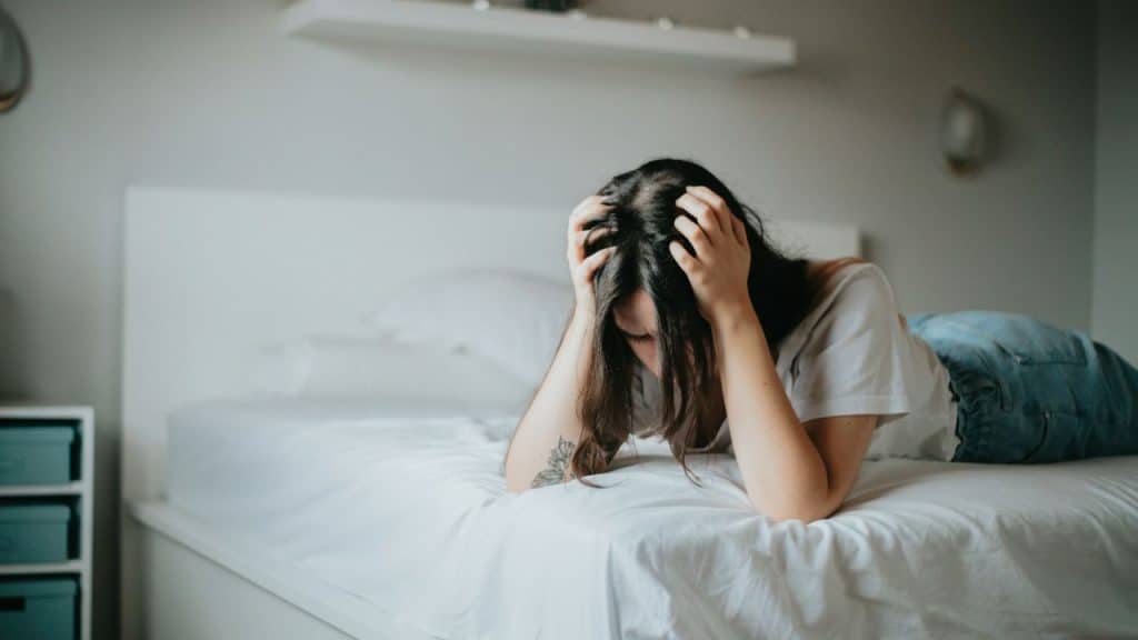 Woman lies face down on a bed, holding her head with both hands in distress.