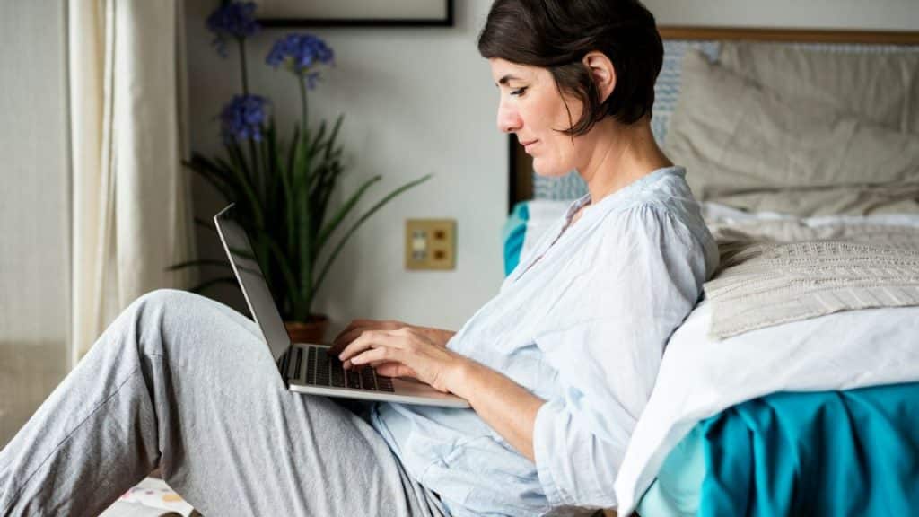Woman sits on the floor leaning against a bed, typing on a laptop.