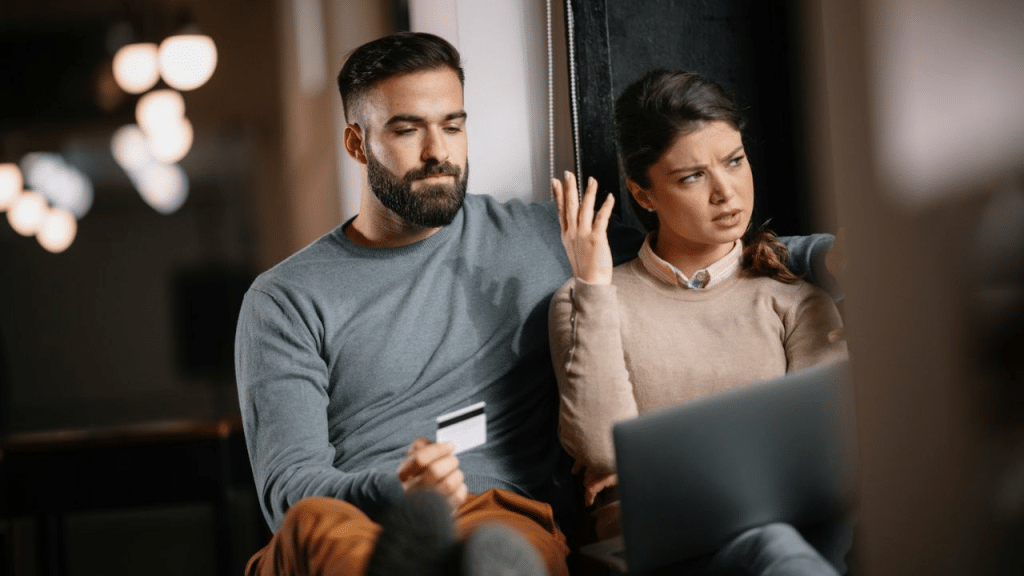 A woman looks annoyed as a man holds a credit card and gestures with one hand.