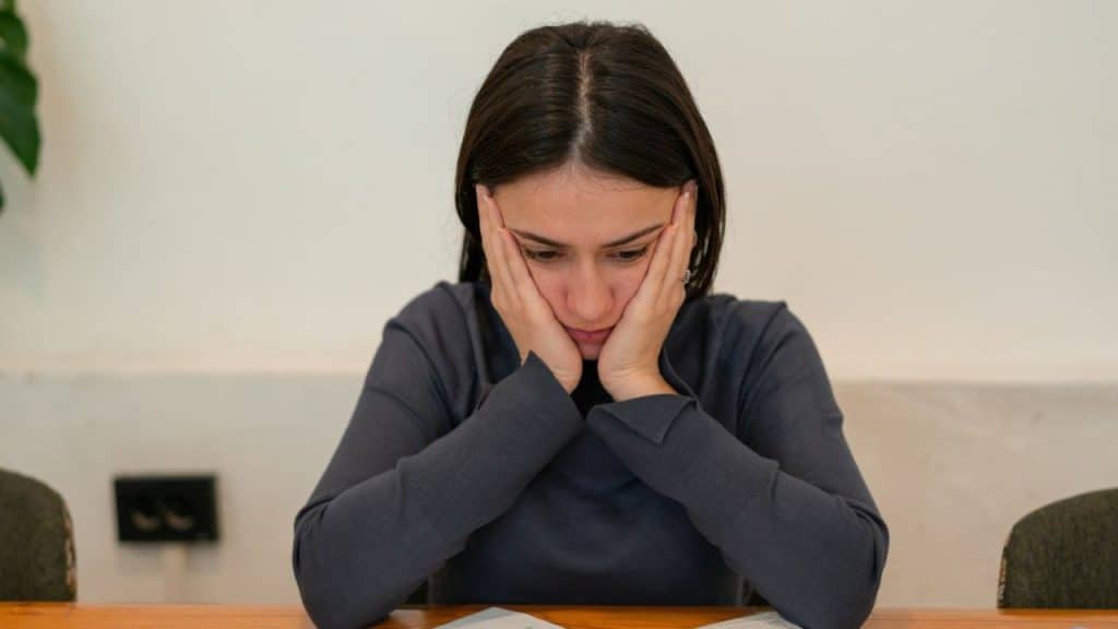 A stressed woman sits at a table with her head in her hands.