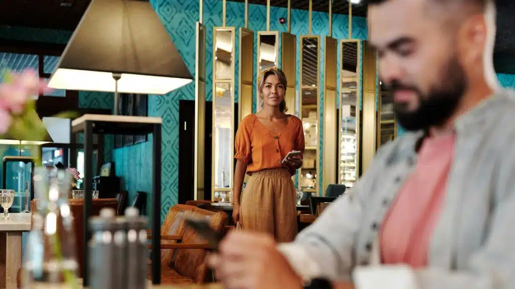 A woman in an orange blouse stands in a restaurant while a man looks at his phone.