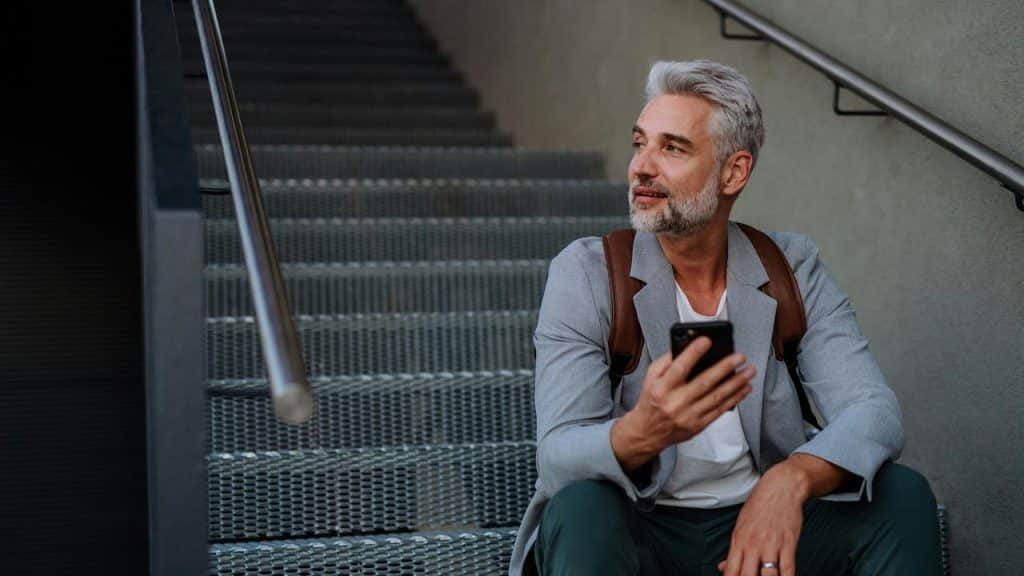 A man with gray hair sits on outdoor steps holding a phone and looking to the side.