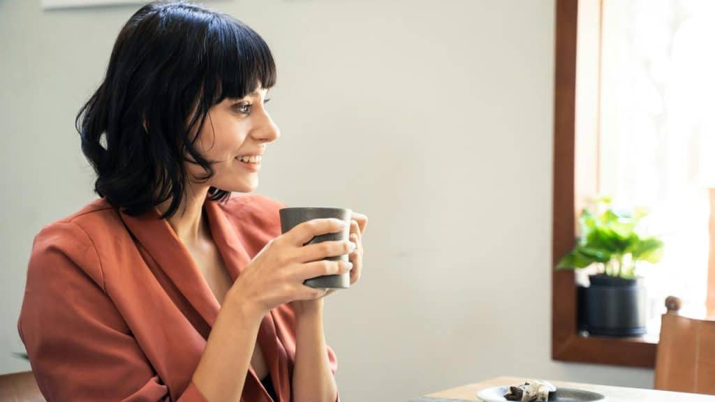 A woman in a rust-colored jacket holds a cup and smiles while sitting at a table.