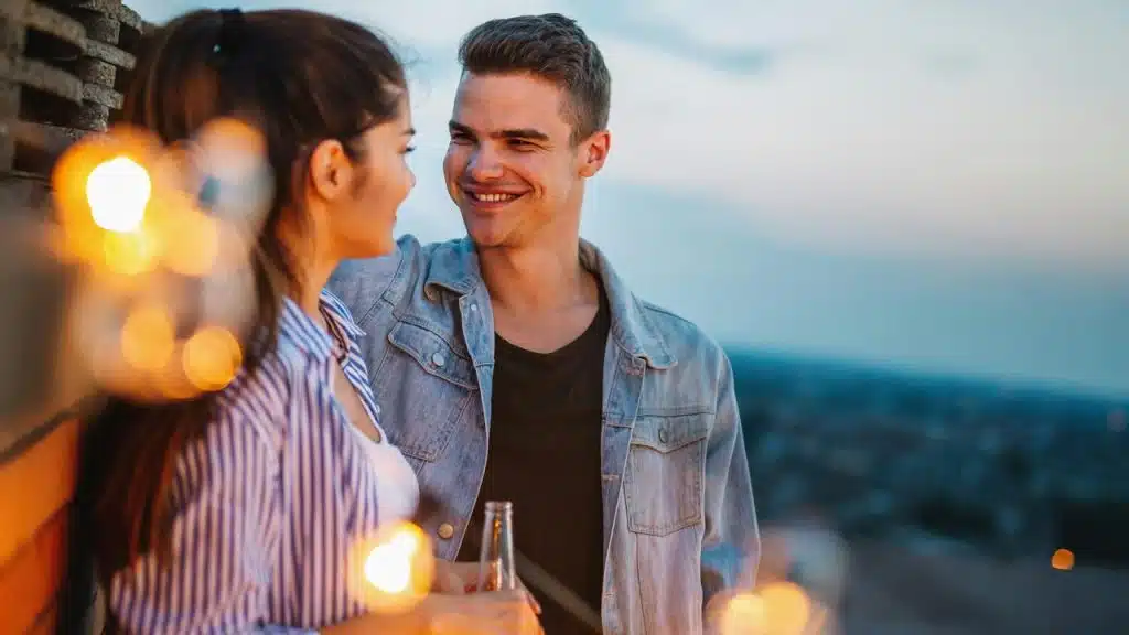 A young couple stands outdoors with drinks, smiling at each other in the evening light.