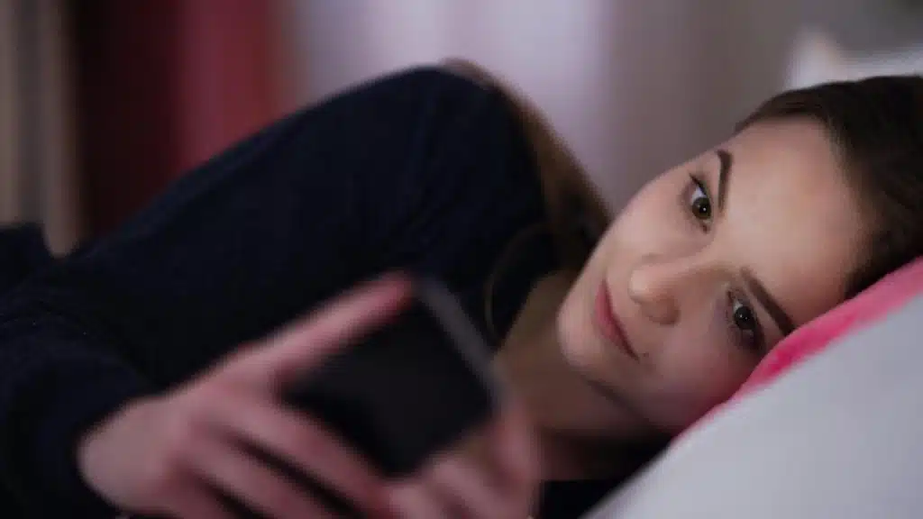 A young woman lies on a bed holding her smartphone while looking ahead.