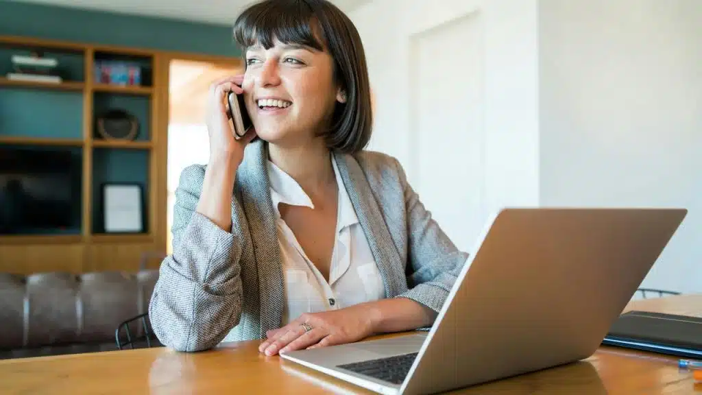 A woman in a blazer smiles while talking on the phone at a desk with a laptop.