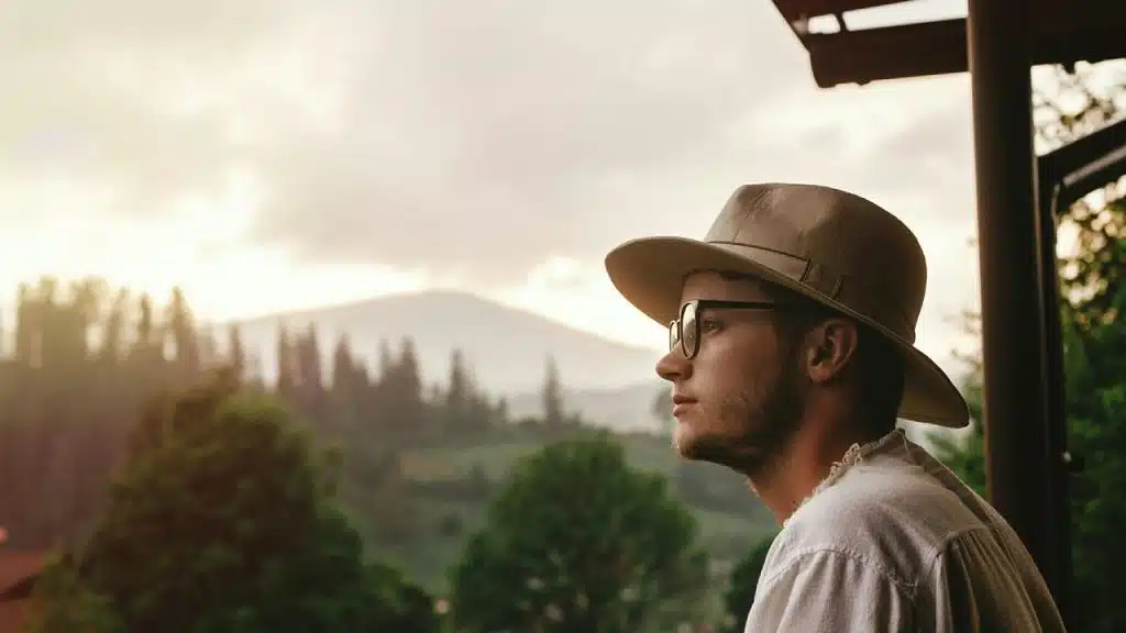 A man in glasses and a wide-brimmed hat looks out at a scenic landscape during sunset.