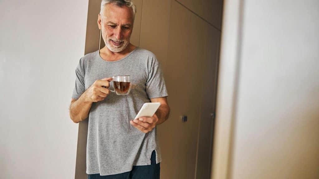 A man with gray hair holding a cup of tea while looking at his phone.