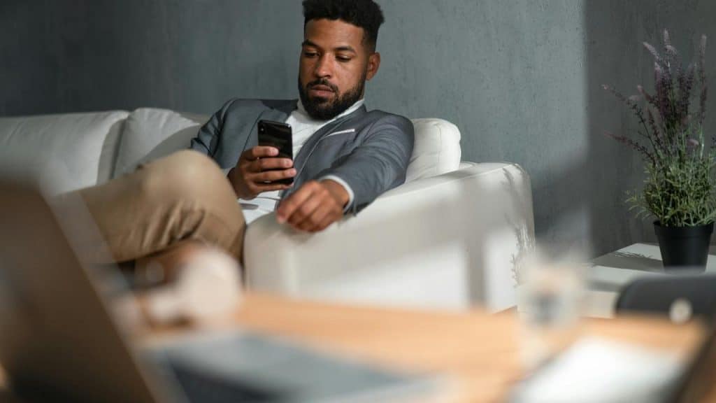 A man in a blazer sitting on a couch while using his phone.