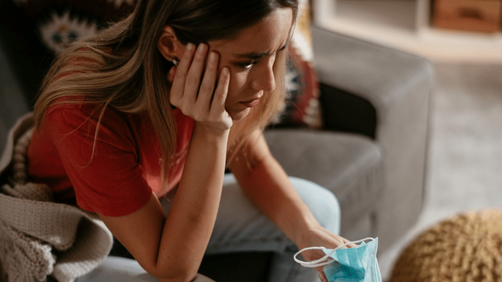 A woman sits on a sofa, looking upset, and holding a face mask.
