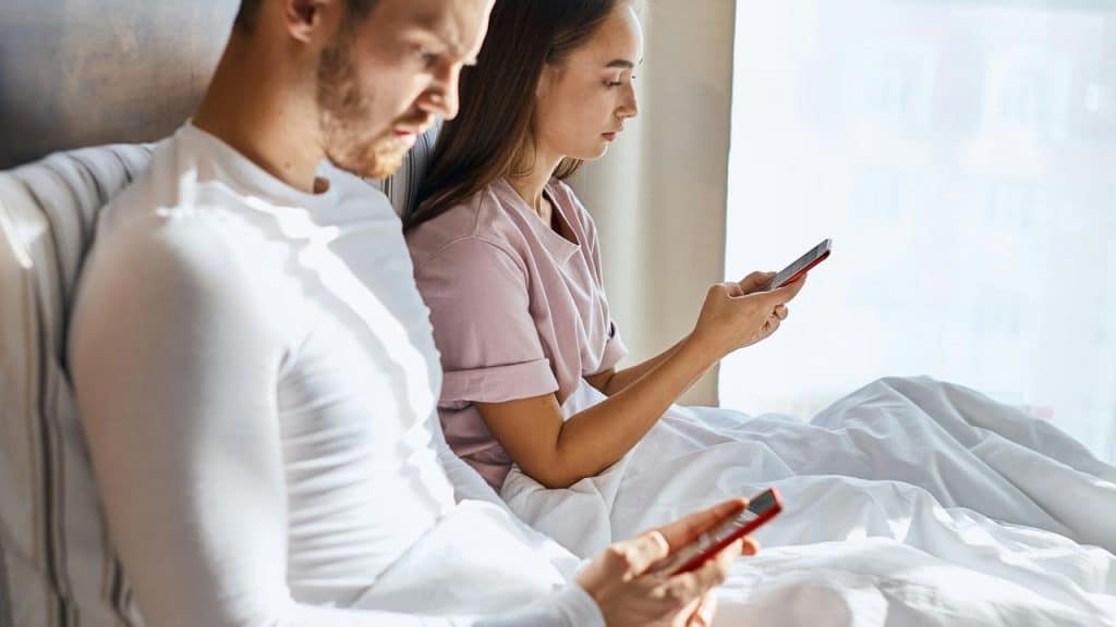 A man and woman sitting in bed while looking at their phones.