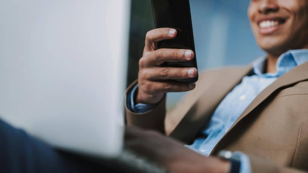 A man in a blazer smiling while holding a smartphone near a laptop.