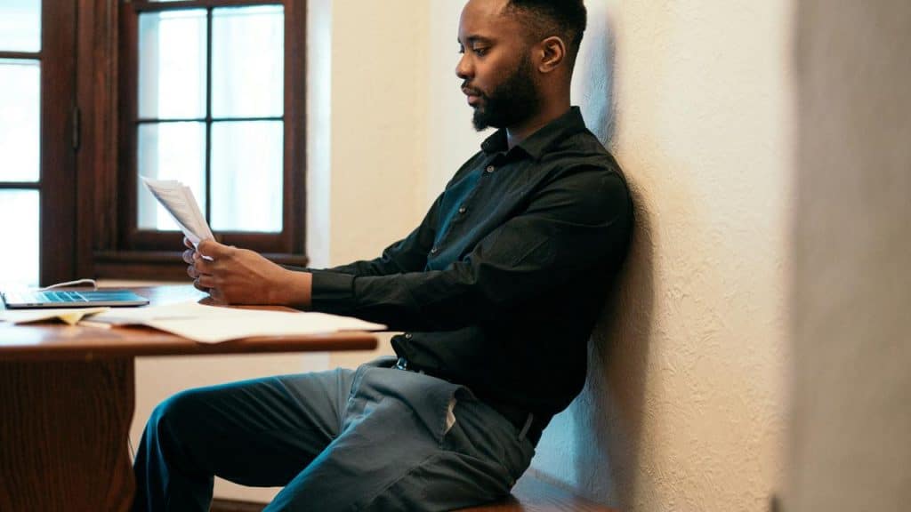 A man in a black shirt sitting by a desk reading papers with a laptop nearby.
