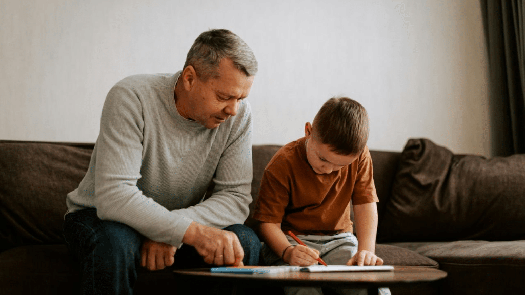 A dad and his young son sit on a couch, with the son drawing.