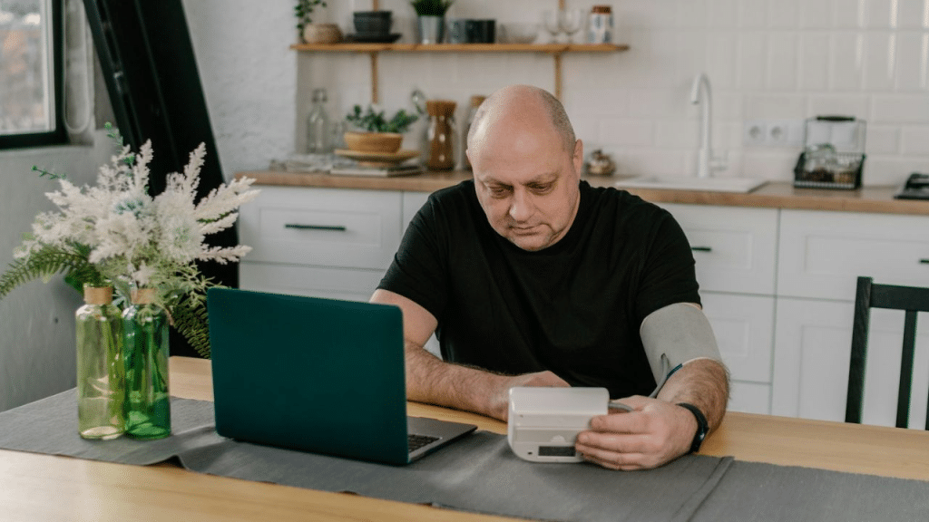A balding man checks his blood pressure at a table with a laptop.