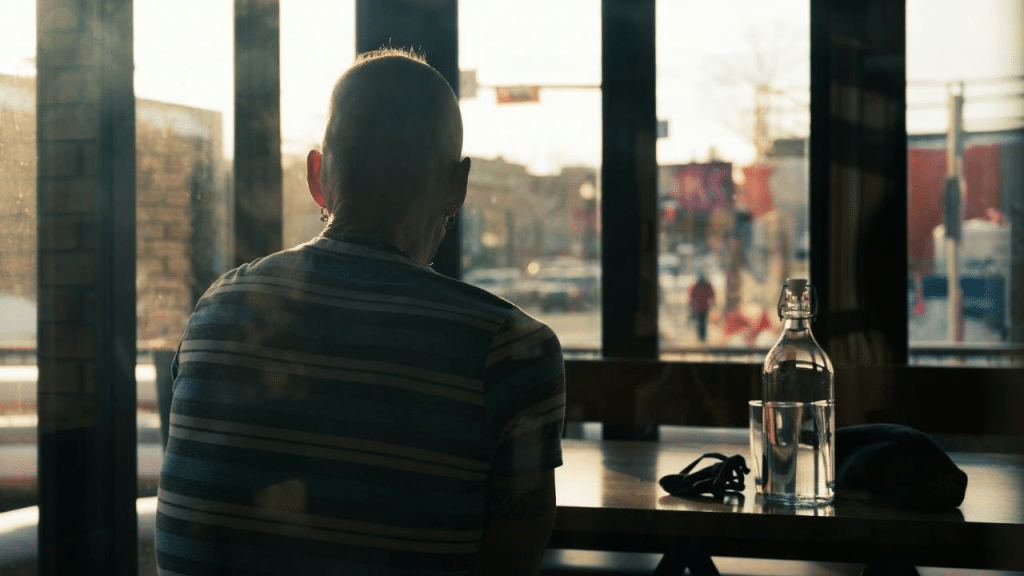 A man with a shaved head sits alone at a table with a glass bottle.