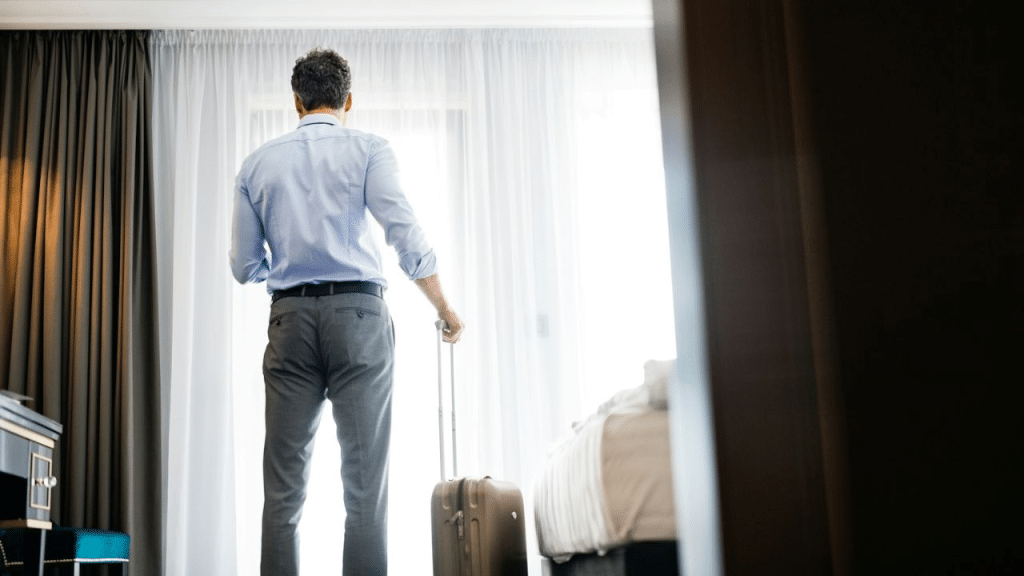 A man in a button-down shirt stands with a suitcase, looking out a window.