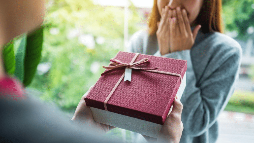 A person presents a wrapped gift to another person who is covering their mouth.