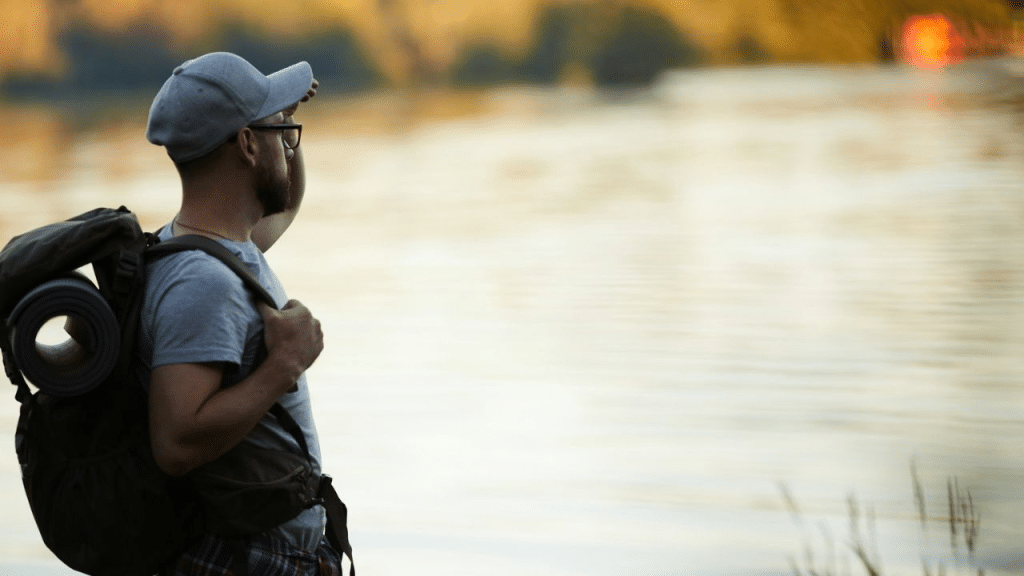 A man with a backpack and a hat looks out at a river.
