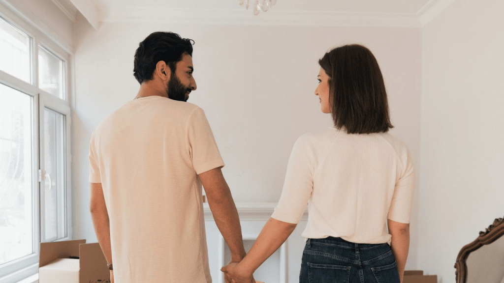 A couple stands in an empty room, holding hands and looking at each other.