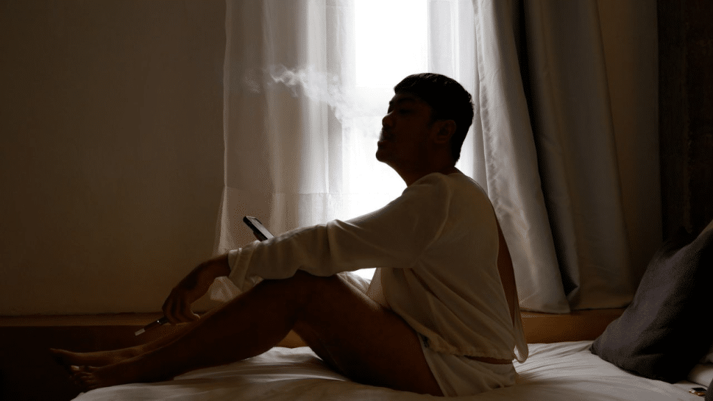 A silhouetted person sits on a bed, looking up and exhaling smoke.