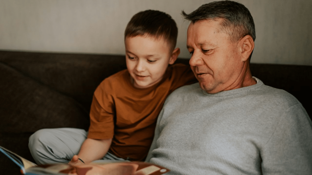 A grandfather and his grandson are sitting on a couch, looking at a book together.