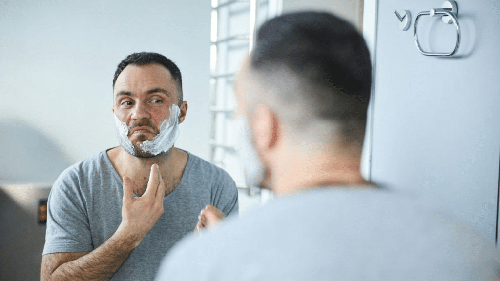 A man with shaving cream on his face looks in a mirror.