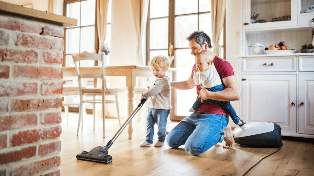 A man holds a baby while a toddler vacuums the floor.