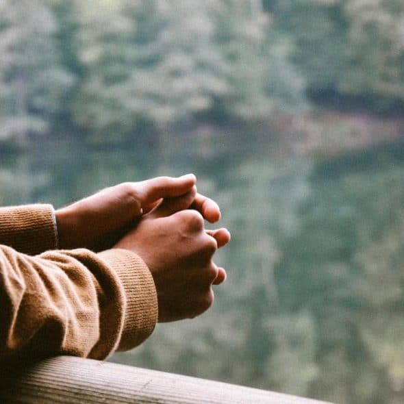 A person resting their clasped hands on a wooden railing while looking out at a lake and trees.