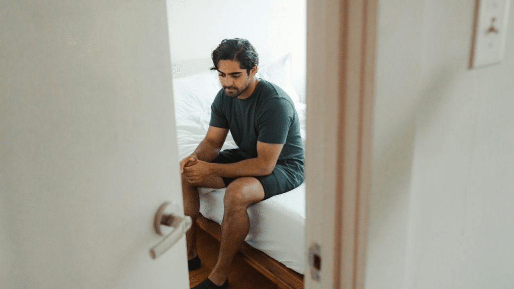 A man sits alone on the edge of a bed, looking down, seen through a doorway.