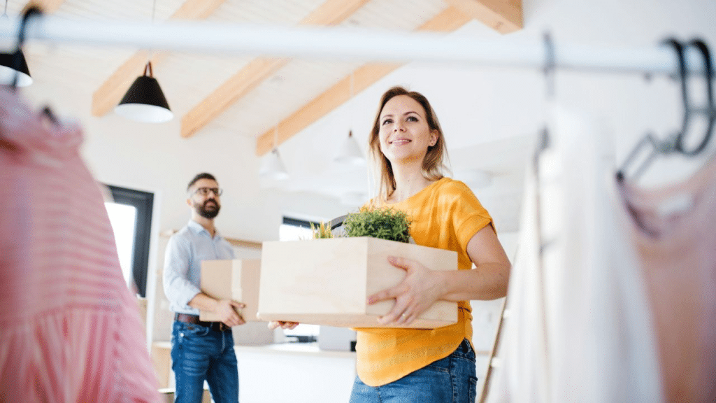 A woman carries a wooden box with a plant while a man in the background carries a cardboard box.