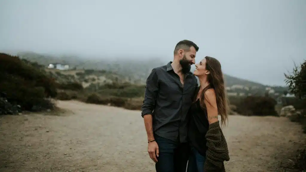 A couple standing close together on a dirt path, smiling at each other.