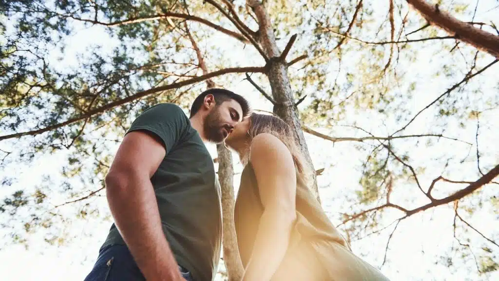 A couple standing close under tall trees, about to kiss.