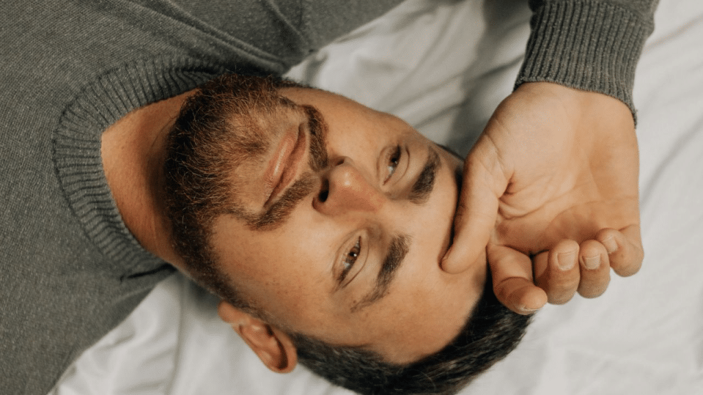 A bearded man in a gray sweater lies on a white bed, resting his hand on his head.
